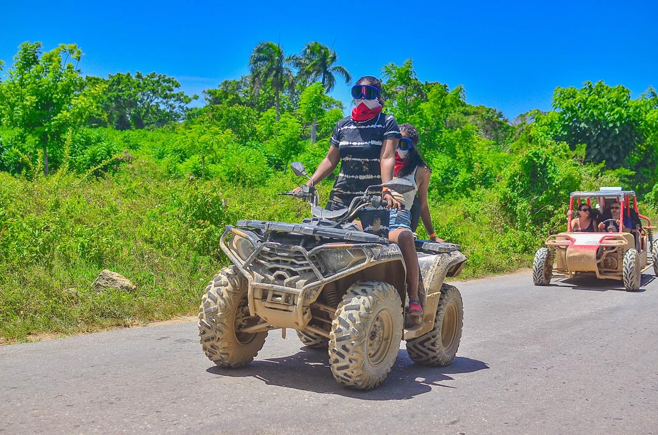 Two travellers on a quad bike on a road through greenery, with a buggy behind